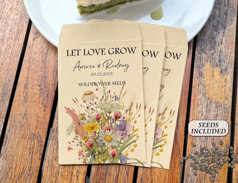 May include: Three kraft paper seed packets with floral illustrations and the text "LET LOVE GROW Aurora & Rodney 05.22.2025 WILDFLOWER SEEDS." A small sign reads "SEEDS INCLUDED." A scattering of seeds is visible.