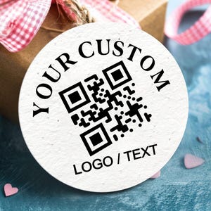 May include: A round white tag with the words "YOUR CUSTOM" and "LOGO / TEXT" printed in black. A QR code is in the center. The tag is tied to a brown gift box with a pink and white checkered ribbon.
