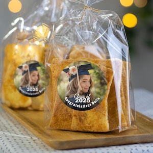 K&ouml;nnte beinhalten: Transparente Plastikt&uuml;ten gef&uuml;llt mit knusprigen, goldbraunen Brotst&auml;bchen. Jede T&uuml;te hat einen runden Aufkleber mit einem Foto einer Absolventin in Kappe und Robe, dem Text "Class of 2025" und den Worten "Congratulations Bella".