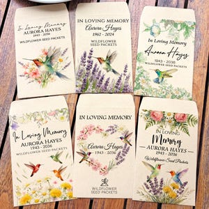 May include: Six wildflower seed packets with floral and hummingbird illustrations. Each packet reads "In Loving Memory" and includes a name and dates. The packets are beige with various floral designs and hummingbird images.