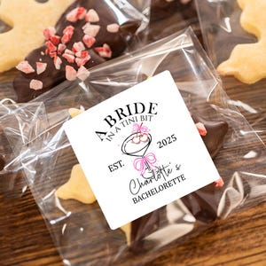 May include: A white sticker with black text that reads "A Bride In A Tini Bit Est. 2025 Charlotte's Bachelorette" with a pink and black illustration of a martini glass with cherries. The sticker is on a clear plastic bag with a chocolate-covered cookie inside.