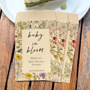 May include: Four seed packets with floral designs and the text "baby in bloom" are arranged on a wooden table. The packets feature daisies, wildflowers, and the text "Madison's Baby Shower 05.26.2025 WILDFLOWER SEEDS". A white plate with a slice of cake is in the background.