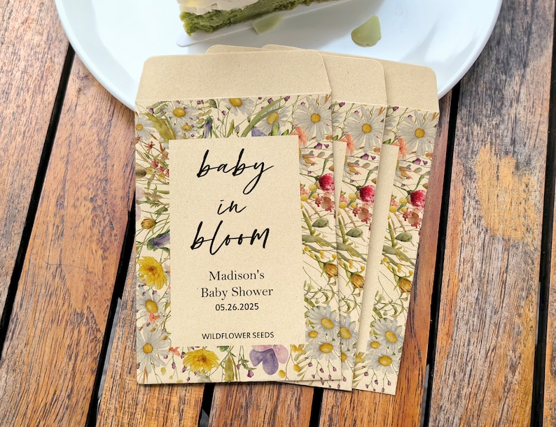 May include: Four seed packets with floral designs and the text "baby in bloom" are arranged on a wooden table. The packets feature daisies, wildflowers, and the text "Madison's Baby Shower 05.26.2025 WILDFLOWER SEEDS". A white plate with a slice of cake is in the background.