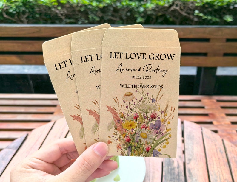 May include: Three kraft paper seed packets with the text "LET LOVE GROW" and "Aurora & Rodney 05.22.2025 WILDFLOWER SEEDS." Each packet features a watercolor floral design with daisies, lavender, and other wildflowers. The packets are held in a person's hand.