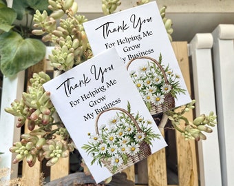 May include: Two white cards with the text "Thank You For Helping Me Grow My Business" in black script. Each card features a watercolor illustration of a basket overflowing with white daisies. The cards are surrounded by green and beige succulents.