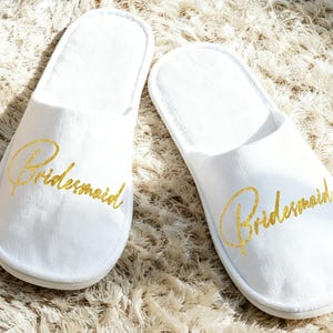 以下が含まれることがあります： 「Bridesmaid」の文字が金色の筆記体で書かれた、白いオープントゥのスリッパのペア。 スリッパは柔らかい素材でできており、明るい色のテクスチャのある表面に置かれています。