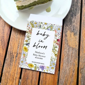 May include: A seed packet with the text "baby in bloom" and "Madison's Baby Shower 05.26.2025." The packet is decorated with a floral design of white, yellow, and purple wildflowers. A slice of green cake sits on a white plate.