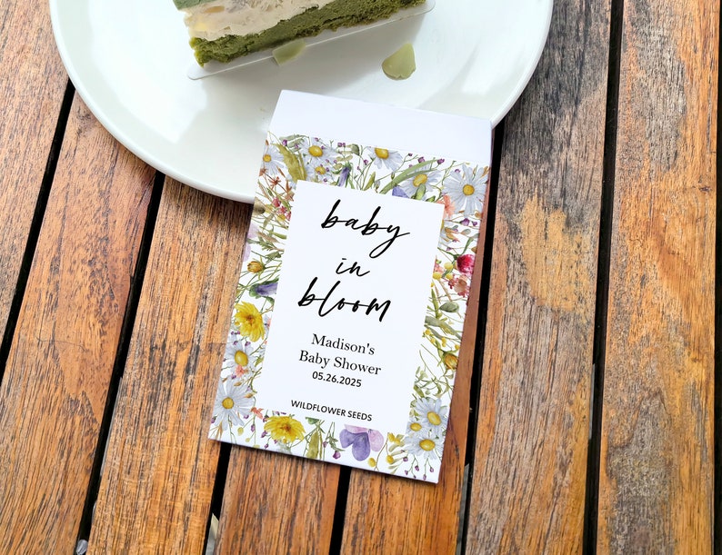 May include: A seed packet with the text "baby in bloom" and "Madison's Baby Shower 05.26.2025." The packet is decorated with a floral design of white, yellow, and purple wildflowers. A slice of green cake sits on a white plate.