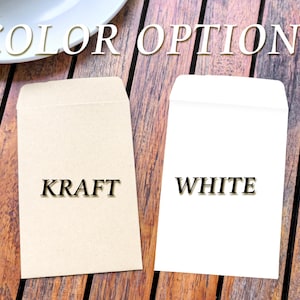 May include: Two small envelopes, one kraft and one white, are displayed on a wooden surface. The word "COLOR OPTIONS" is written above the envelopes. The kraft envelope has the word "KRAFT" printed on it, and the white envelope has the word "WHITE" printed on it.