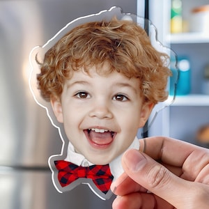 K&ouml;nnte beinhalten: Ein klarer Acrylmagnet mit einem l&auml;chelnden Kind mit lockigem rotem Haar und einer rot-schwarz karierten Fliege. Der Magnet wird vor einem K&uuml;hlschrank aus Edelstahl gehalten.