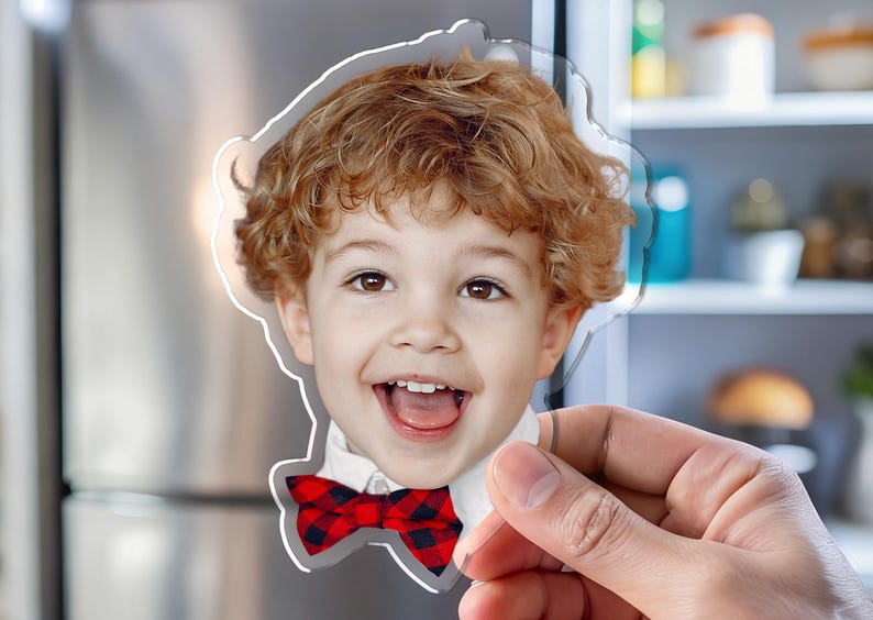 K&ouml;nnte beinhalten: Ein klarer Acrylmagnet mit einem l&auml;chelnden Kind mit lockigem rotem Haar und einer rot-schwarz karierten Fliege. Der Magnet wird vor einem K&uuml;hlschrank aus Edelstahl gehalten.