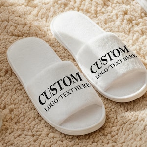 Könnte beinhalten: Ein Paar weiße offene Hausschuhe mit dem Wort "CUSTOM" und "LOGO/TEXT HERE" in Schwarz aufgedruckt. Die Hausschuhe sind aus weichem, plüschigem Material und liegen auf einem beigen Teppich.