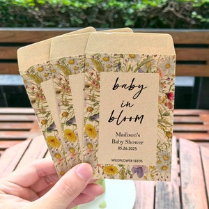 May include: Four seed packet favors with a floral design and the text "baby in bloom" for a baby shower. The packets are tan with a floral border and the text "Madison's Baby Shower 05.26.2025 Wildflower Seeds".
