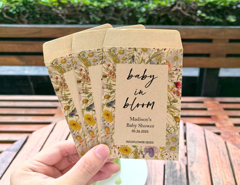 May include: Four seed packet favors with a floral design and the text "baby in bloom" for a baby shower. The packets are tan with a floral border and the text "Madison's Baby Shower 05.26.2025 Wildflower Seeds".