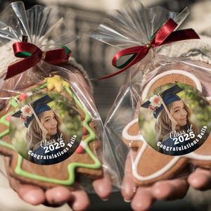 K&ouml;nnte beinhalten: Zwei dekorierte Lebkuchen in durchsichtigen Plastikt&uuml;ten, die mit einem roten Band zusammengebunden sind. Die Kekse sind in Form eines Weihnachtsbaums und eines Lebkuchenmanns geformt. Jeder Keks hat einen runden Aufkleber mit einem Foto eines Absolventen, der eine M&uuml;tze und eine Robe tr&auml;gt, und dem Text "Class of 2025 Congratulations Bella".