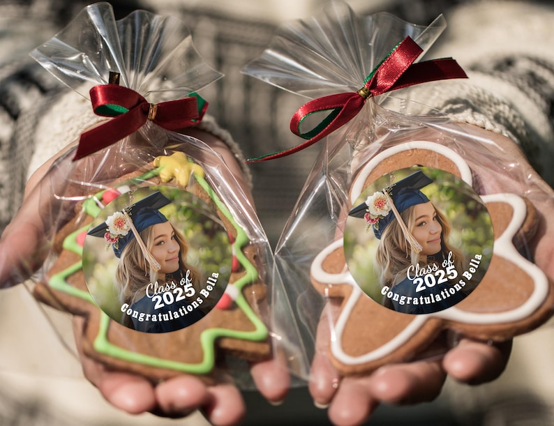 K&ouml;nnte beinhalten: Zwei dekorierte Lebkuchen in durchsichtigen Plastikt&uuml;ten, die mit einem roten Band zusammengebunden sind. Die Kekse sind in Form eines Weihnachtsbaums und eines Lebkuchenmanns geformt. Jeder Keks hat einen runden Aufkleber mit einem Foto eines Absolventen, der eine M&uuml;tze und eine Robe tr&auml;gt, und dem Text "Class of 2025 Congratulations Bella".