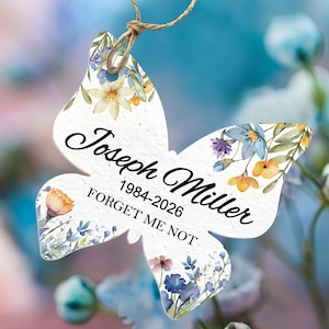 May include: A butterfly-shaped memorial ornament with floral watercolour designs in blue, yellow, and orange. The ornament has the name "Joseph Miller" and dates "1984-2026" with the words "Forget Me Not". It is suspended by a twine string.