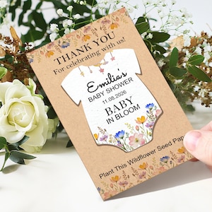 May include: A baby shower invitation card with a floral design. The card features the text "THANK YOU" and "Emilia's BABY SHOWER 11.08.2026 BABY IN BLOOM". The card is decorated with colorful flowers and a baby onesie illustration.