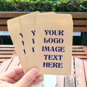 Op de afbeelding: Een hand houdt drie kleine, lichtbruine papieren enveloppen vast. Op de voorste envelop staat de tekst "YOUR LOGO IMAGE TEXT HERE" in donkerblauw gedrukt. De enveloppen liggen op een houten tafel.
