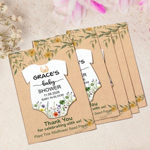 May include: A stack of baby shower invitations. Each invitation features a white onesie shape with the text "GRACE'S baby SHOWER 11.08.2026 BABY IN BLOOM". The invitations are on a tan background with floral accents.