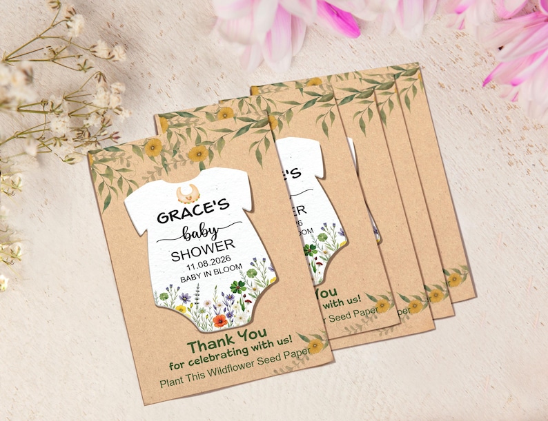 May include: A stack of baby shower invitations. Each invitation features a white onesie shape with the text "GRACE'S baby SHOWER 11.08.2026 BABY IN BLOOM". The invitations are on a tan background with floral accents.