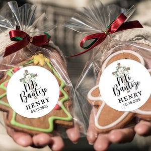 May include: Two gingerbread cookies decorated with white frosting and a round sticker. The sticker has a white background with a green floral design, a wooden cross, and the text "Mi Bautizo HENRY 05.18.2025".