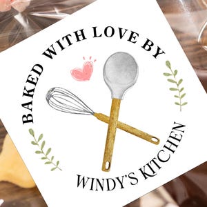 May include: A square sticker with the text "Baked with Love by Windy's Kitchen" encircling a whisk and spoon. A pink heart and green sprigs accent the design. The sticker is on a clear cellophane bag.