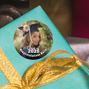 K&ouml;nnte beinhalten: Ein runder Abschlussaufkleber mit einem Foto einer jungen Frau in einer Abschlusscap und -robe. Der Aufkleber tr&auml;gt den Text "Class of 2025" und "Congratulations Bella".
