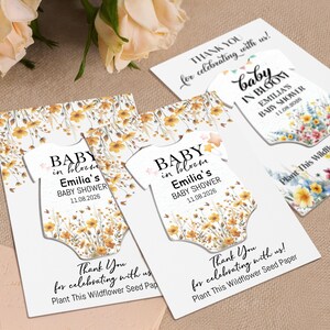 May include: Baby shower seed paper cards with a floral design. The cards feature a baby onesie shape with the text "Baby in Bloom" and event details. The cards are white with a floral pattern and the text "Thank You for celebrating with us!"