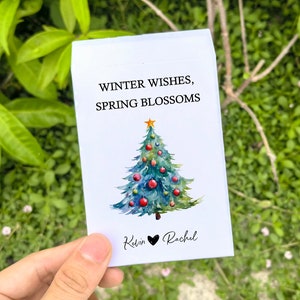 May include: A white card with the text "WINTER WISHES, SPRING BLOSSOMS" above a watercolor Christmas tree decorated with red ornaments and a gold star. The names "Kevin" and "Rachel" are written below the tree.