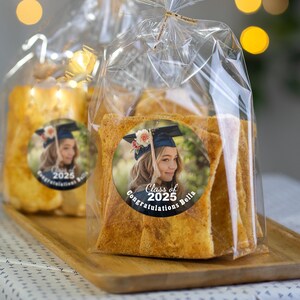 K&ouml;nnte beinhalten: Transparente Plastikt&uuml;ten gef&uuml;llt mit knusprigen, goldbraunen Brotst&auml;bchen. Jede T&uuml;te hat einen runden Aufkleber mit einem Foto einer Absolventin in Kappe und Robe, dem Text "Class of 2025" und den Worten "Congratulations Bella".