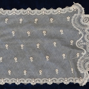May include: A white lace fabric with a delicate floral pattern. The lace has a scalloped edge with intricate designs.