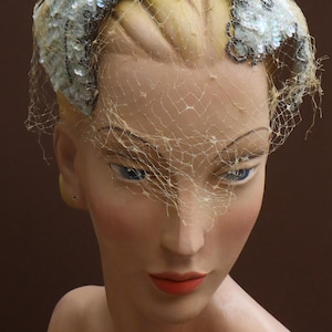 Può includere: Un cappello bianco e argento con paillettes e velo. Il cappello è indossato da un manichino con un incarnato marrone chiaro e rossetto rosso.