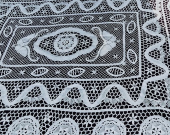 Könnte beinhalten: Weiße Häkeltischdecke mit einem zarten Blumenmuster und filigranen Spitzenarbeiten. Die Tischdecke hat ein zentrales quadratisches Design mit einem kreisförmigen Motiv und einen Rand aus sich wiederholenden ovalen Formen.