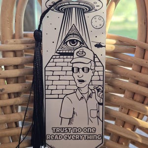 May include: A white bookmark with a black illustration of a pyramid with an eye, a UFO, and a man wearing sunglasses. The text "TRUST NO ONE READ EVERYTHING" is printed at the bottom. A black tassel hangs from the top.