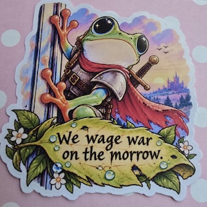 May include: A die-cut sticker featuring a green frog dressed as a warrior with a sword and red cape, climbing a tree. Below the frog is a large yellow leaf with the text "We wage war on the morrow." The background shows a sunset landscape.