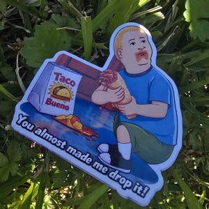 May include: A sticker featuring a cartoon image of a boy holding a burrito, with a spilled box of "Taco Bueno" and the text "You almost made me drop it!". The boy is wearing a blue shirt and green shorts.