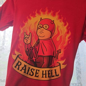 May include: A bright red short-sleeved t-shirt featuring a graphic of a cartoon character wearing a red devil mask and cape, surrounded by orange and yellow flames. The character holds a trident and makes a hand gesture. A banner reads RAISE HELL.