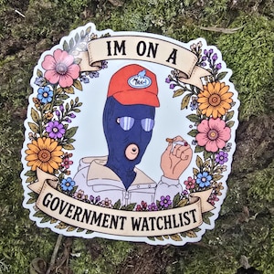 May include: A die-cut sticker featuring a person in a blue balaclava, orange cap, and sunglasses, holding a cigarette. The text reads IM ON A GOVERNMENT WATCHLIST. The figure is framed by colourful flowers in shades of pink, orange, and purple.