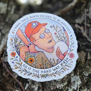 Puede incluir: Una pegatina redonda con una ilustración de dibujos animados de un hombre con gafas y una gorra roja, siendo tirado por una mano. La pegatina tiene el texto "I HAVE ONCE AGAIN DECIDED TO BE DIFFICULT. DO IT THE HARD WAY."