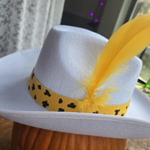May include: A white fedora-style hat with a yellow band featuring black spots. A large yellow feather is tucked into the hatband. The hat is resting on a pumpkin, suggesting a fall or Halloween theme.