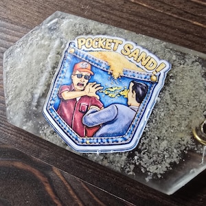 Könnte beinhalten: Ein klarer, sechseckiger Schlüsselanhänger mit einer Cartoon-Grafik eines Mannes, der Sand wirft. Die Grafik lautet "POCKET SAND!" und zeigt einen Mann in einem roten Hemd und Sonnenbrille. Der Schlüsselanhänger hat einen goldfarbenen Ring.