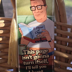 May include: Bookmark featuring a cartoon image of a man reading a book titled "A Dinner of Onions." The bookmark has a tassel and the text "This page isn't gonna turn itself, I'll tell you h'wat."