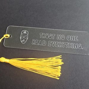 May include: A clear acrylic bookmark with the text "TRUST NO ONE READ EVERYTHING." A yellow tassel is attached to the top. The bookmark features a line drawing of a person wearing a hat and glasses.