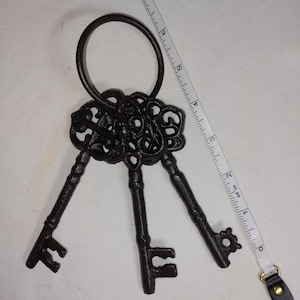 May include: A set of three antique-style, dark brown metal keys with ornate designs, connected by a large ring. A white measuring tape with black markings is placed next to the keys, showing inches. The keys are likely decorative.