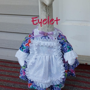 May include: A goose statue wearing a purple floral dress with a white eyelet apron and collar. The dress has butterfly and floral patterns. The word "Eyelet" is written in pink script across the image.