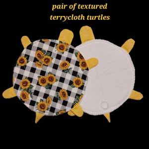 May include: A pair of textured terrycloth turtles with yellow limbs and heads. One turtle features a black and white buffalo check pattern with yellow sunflowers, while the other is solid white terrycloth with a small circular plastic ring attached.