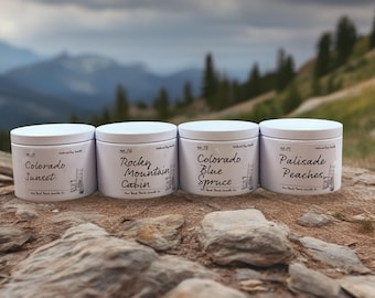 Op de afbeelding: Vier witte kaarsenpotten met deksel met de opschriften "Colorado Sunset", "Rocky Mountain Cabin", "Colorado Blue Spruce" en "Palisade Peaches". De kaarsen staan op een rotsachtig oppervlak met een berglandschap.