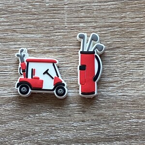Peut inclure: Deux petites breloques décoratives sur le thème du golf. L'une représente une voiturette de golf blanche et rouge avec un sac de golf à l'arrière. L'autre est un sac de golf rouge contenant plusieurs clubs de golf gris à poignées noires.