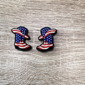 May include: Two small, black cowboy boot charms. Each boot features a design of the American flag, with a blue upper adorned with white stars and red and white stripes. The charms are likely for shoes or accessories.